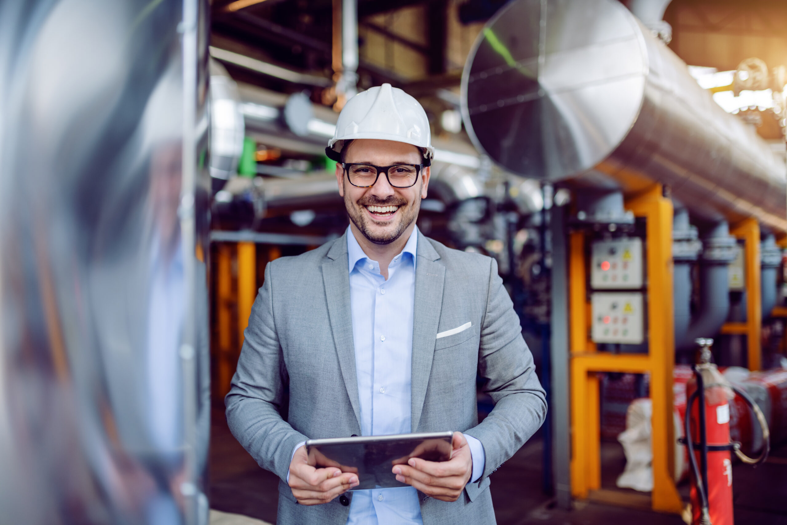 A young man wearing a hard hat while holding a tablet in his hands and smiling